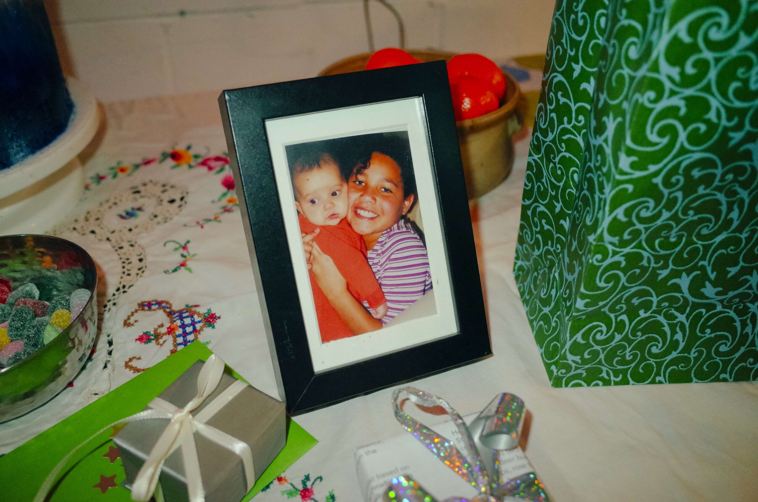 A framed photo of Kyra and Sierra as children sits on a table loaded with party treats and gifts. In the image, Kyra is smiling widely squeezing baby Sierra close to her chest.