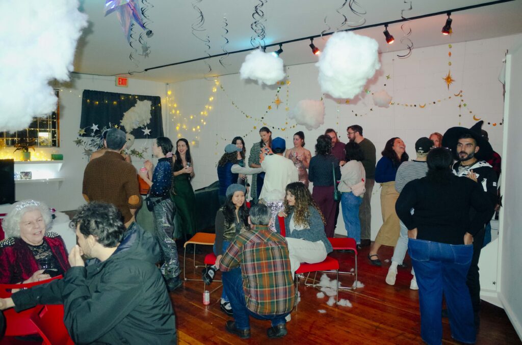 A crowd of partygoers mill about talking and laughing in a room decorated with star streamers, cotton ball clouds and fairy lights. Some people are wearing glittering clothing or spooky costumes.