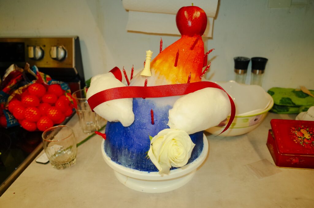 A large teardrop shaped cake iced in a vivid ombre in the colours of sunset rests on a kitchen counter. It is decorated with red birthday candles and various twilight themed objects, including a white rose, a red ribbon, a white queen chess piece, and a red apple.