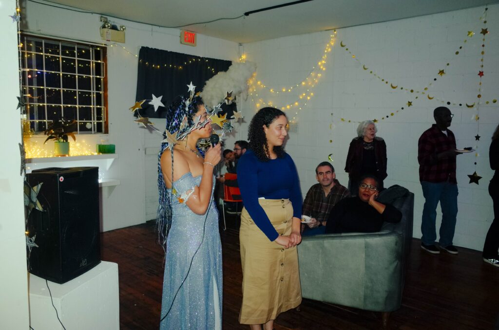 Kyra, wearing a sparkling floor length off the shoulder gown and a crown of silver stars, sings into a microphone. Sierra stands adjacent wearing a khaki skirt and flowing black wig. Partygoers are milling around watching the karaoke set.