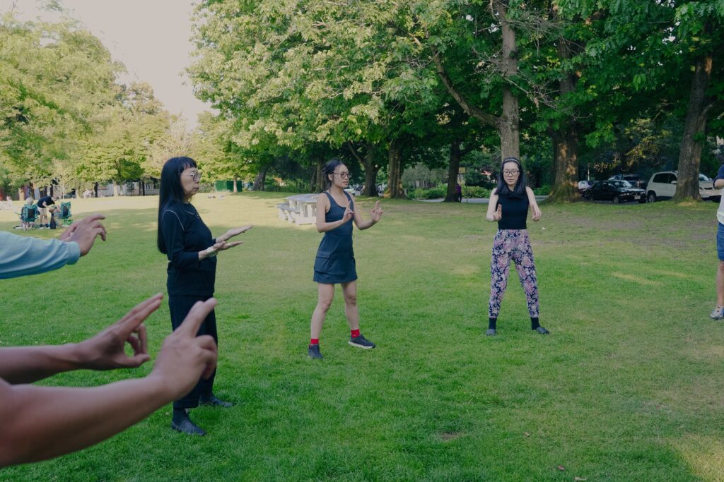 A group of people stand grounded in a wide circle in a grassy park field. Their eyes are closed and their hands are held out in front of their bodies in a mudra like gesture. Breath and movement united in this suspended moment.