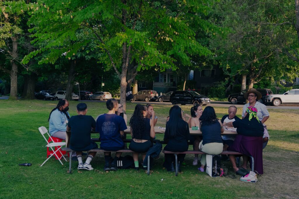Vanessa, wearing a large brim straw hat, speaks to a picnic table full of participants in a sun dappled park field.
