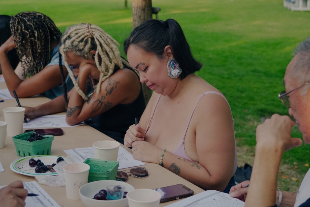 Ramona, wearing large beaded earrings and smiling slightly, looks down at the diagramed page on the picnic table before them. There are other participants on either side of them working on their own pages.