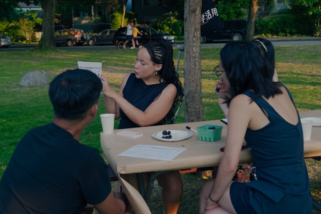 Maya holds up their worksheet page to a group of three participants sitting around a picnic table, munching on fresh blackberries.