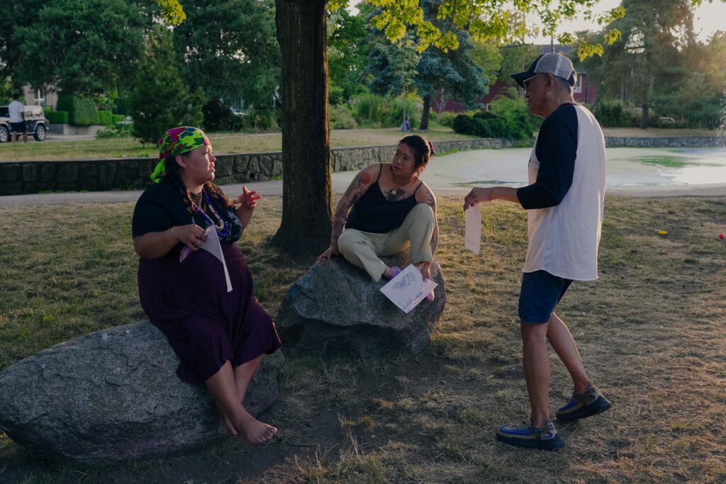 JB the First Lady gestures with their hands while speaking to Beverly perched on a bolder, and Paul standing near by. The evening sun dappling everyones faces and the park around them.