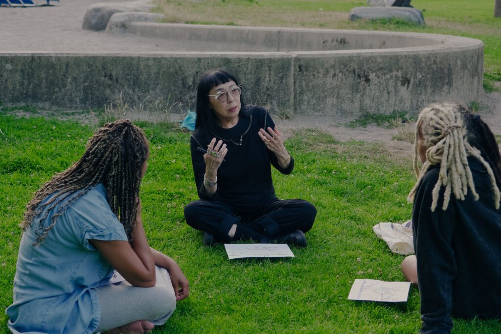 Marilyn sits cross legged on the grass, the light reflecting off her bold white and black cat eye glasses. She is gesturing meaningfully with her hands as she speaks to two participants sitting across from her.