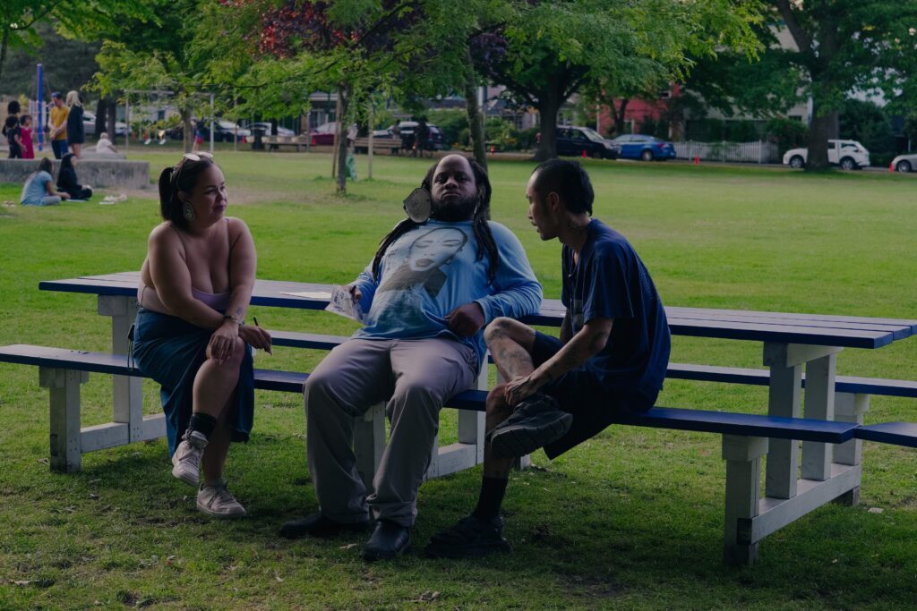 Brandon sits between Ramona and Vinson on one side of a picnic table bench. The three look to be engaged in intense conversation.