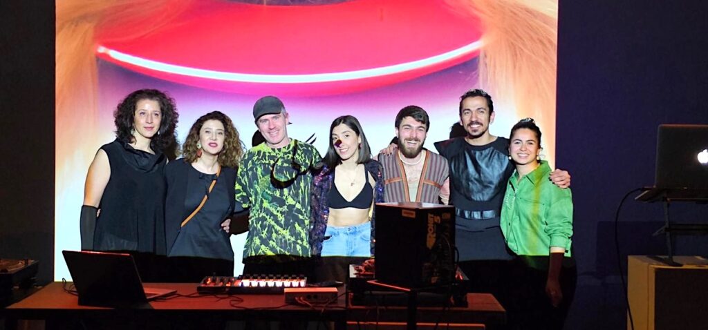 In a darkened room, Shadi Jianfar, Pegah Tabassinejad, Ian Prentice, Saghi Ehteshamzadeh, Parsa Nazeri, Behrad Tehrani, and Kimia Koochakzadeh-Yazdi post for a photo. With arms linked they stand behind a table of sound equipment, their smiling faces are illuminated by the neon colours of the visuals projected onto them and the wall behind.